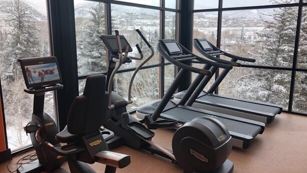 Ein Fitnessraum mit Cardiogeräten und einem Fernsehbildschirm, große Fenster mit Blick auf eine verschneite Landschaft.