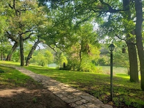 Stroll along the serene park pathway, surrounded by lush greenery and mature trees, leading to a tranquil body of water.
