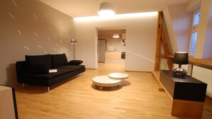 Una sala de estar moderna con un sofá negro, una mesa de centro redonda y un escritorio de madera con una lámpara. Se ve una cocina al fondo.