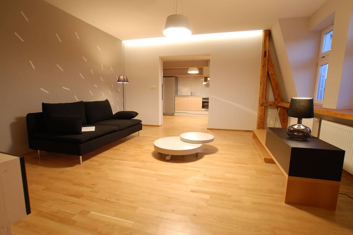A modern living room with a black sofa, a round coffee table, and a wooden desk with a lamp. A kitchen is visible in the background.