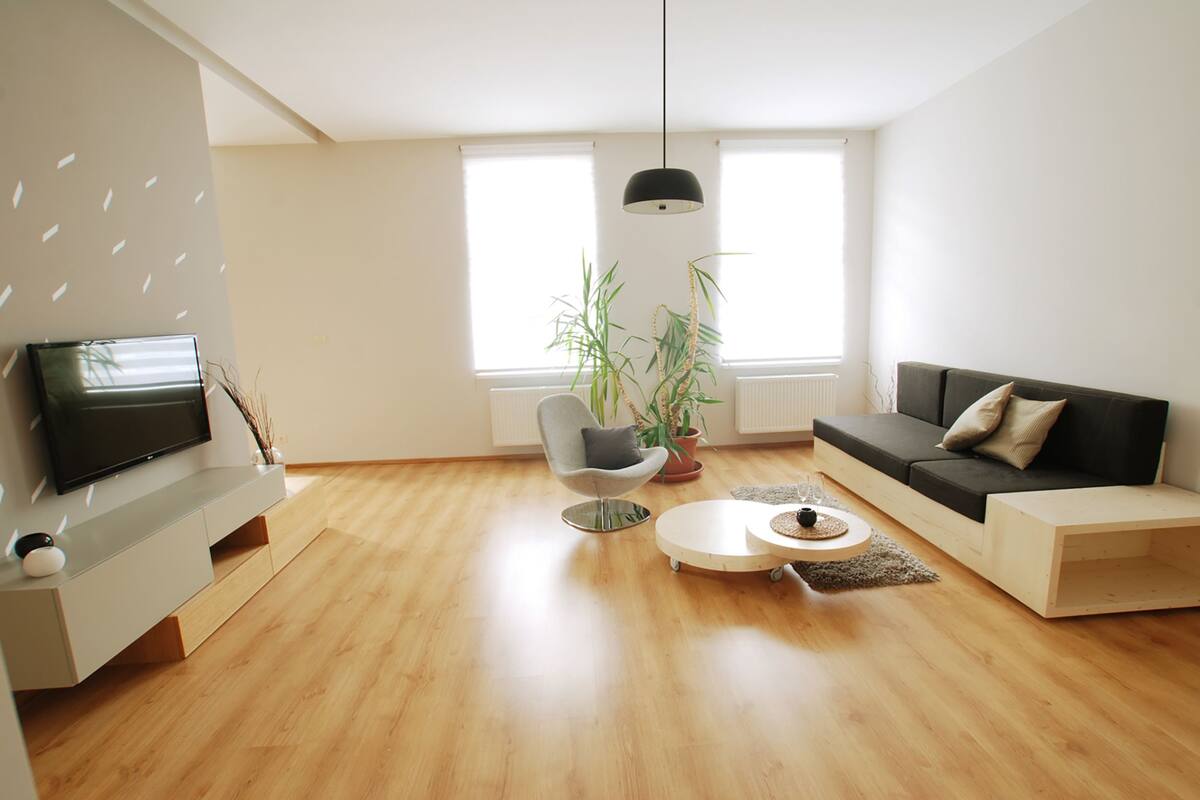 A modern living room with a flat-screen TV, a grey chair, a black sofa, a round coffee table, and a potted plant.