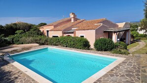 Una piscina rectangular con losetas de piedra alrededor, con una casa de techo con tejas y un cielo azul despejado de fondo.