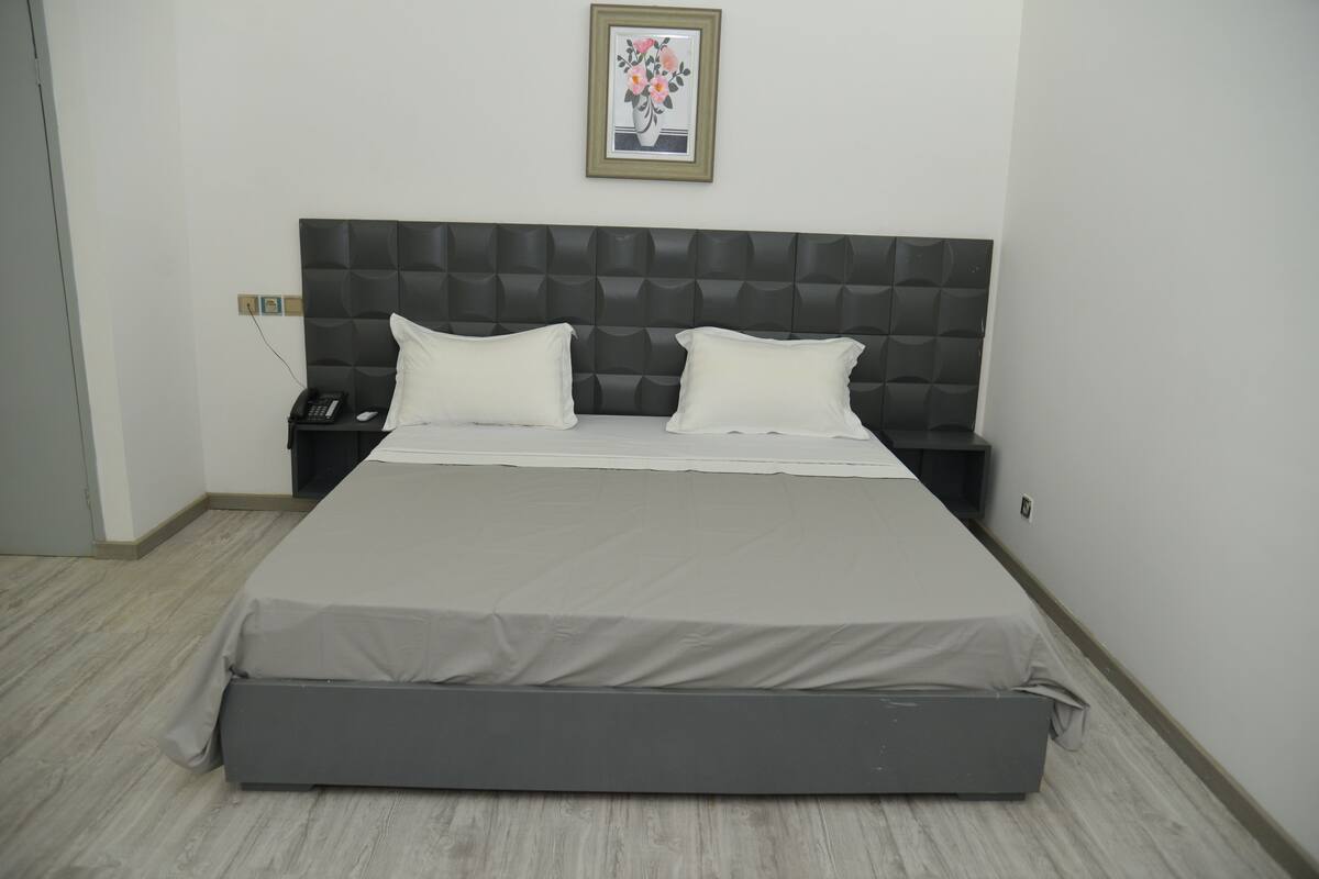 Uma cama bem arrumada com colchão cinza e roupa de cama branca, uma cabeceira escura, uma mesinha de cabeceira com um telefone e um quadro na parede.