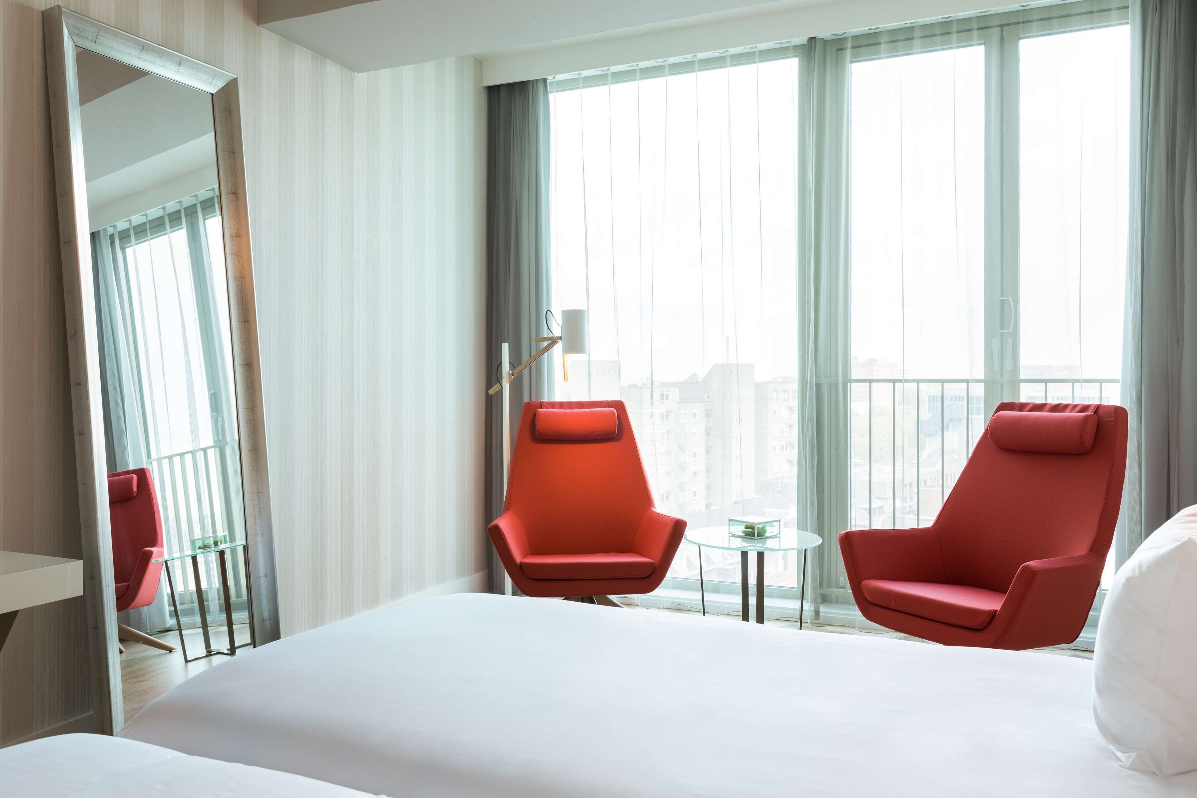 Ett hotellrum med en stor säng, två röda fåtöljer, ett litet bord och ett stort fönster med utsikt över staden.