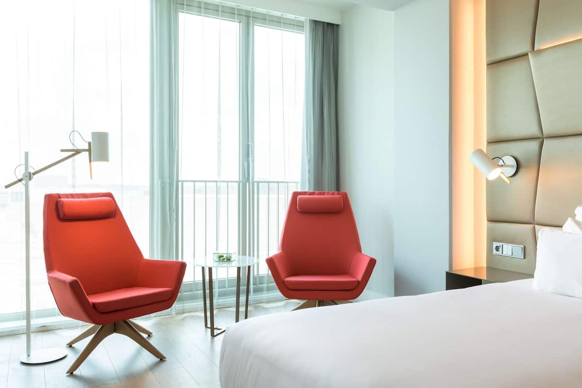 A hotel room with two red chairs, a bed, a bedside table, a lamp, and a large window with curtains.