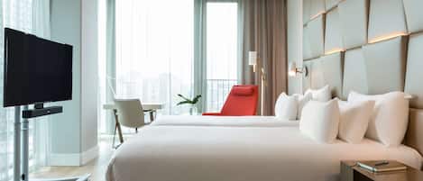 Una habitación de hotel moderna con una cama grande, un televisor sobre un mueble, una silla roja y vistas a la ciudad a través de amplios ventanales.