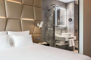 Habitación de hotel moderna con una cama grande, cabina de ducha con paredes de vidrio, tocador con espejo y una lámpara de noche.