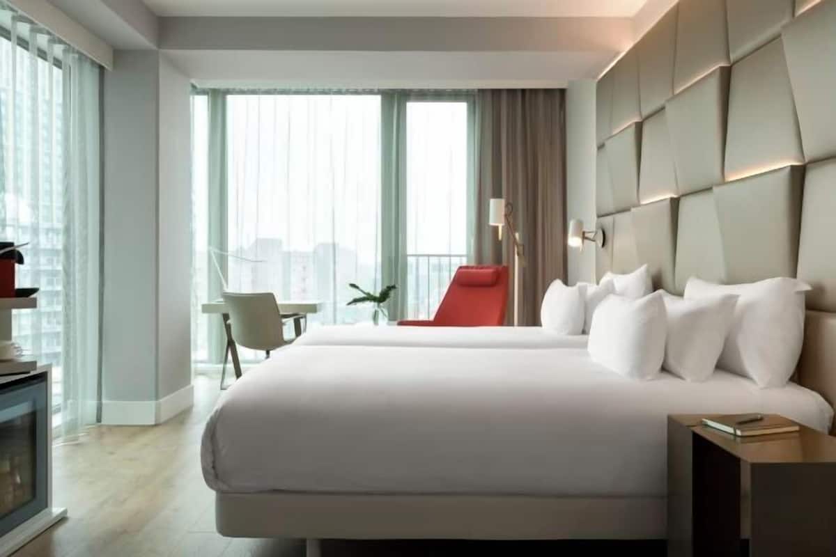 A modern hotel room with a large bed, a red chair, a small table, and a city view.