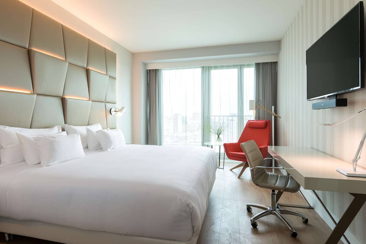 A hotel room with a large bed, a desk with a chair, a flat-screen TV, and a view of the city through large windows.