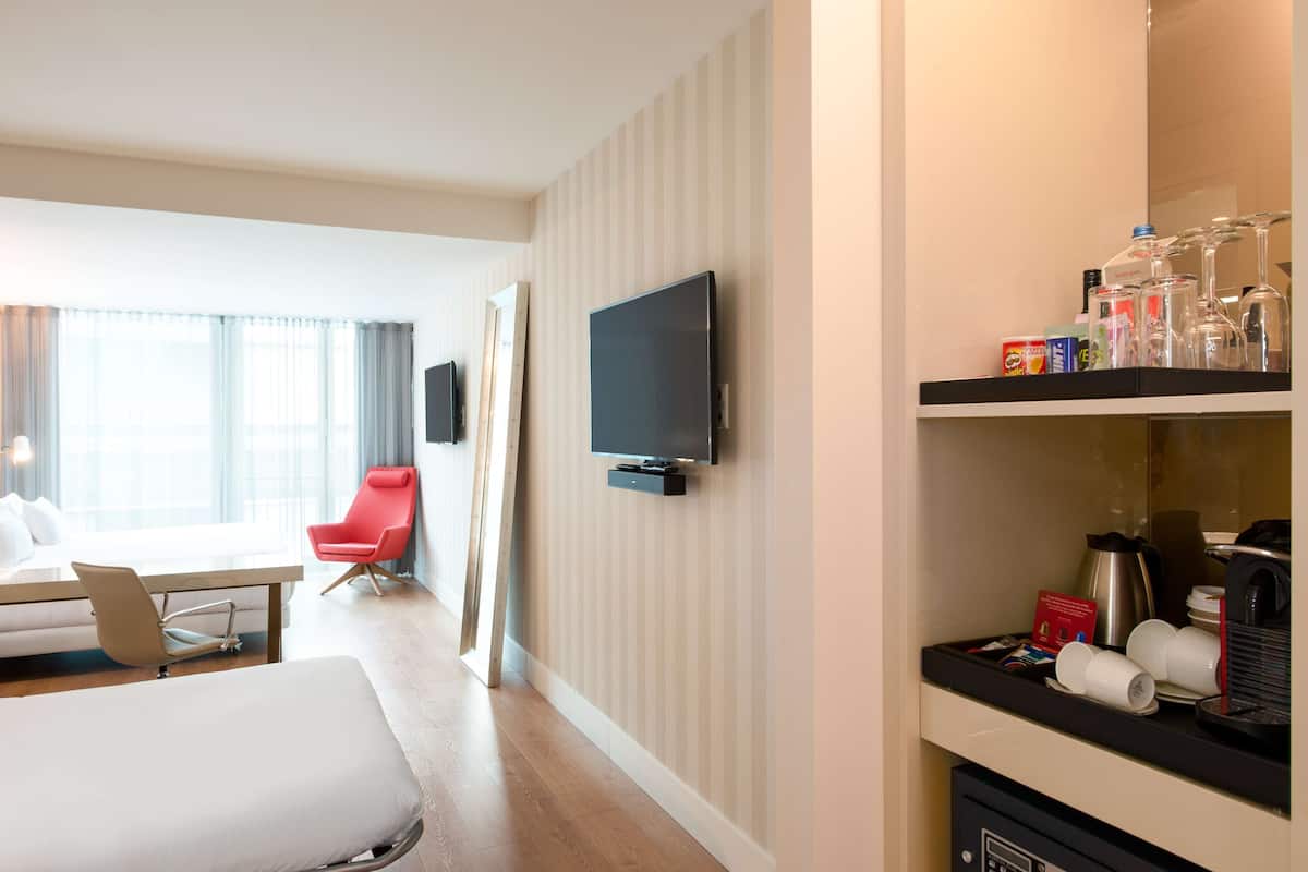 A hotel room with a bed, a red chair, a TV mounted on the wall, and a coffee station with cups and a coffee maker.