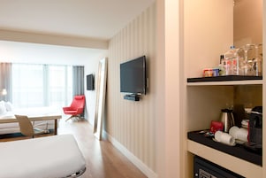 Una habitación de hotel con una cama, una silla roja, un televisor montado en la pared y una zona de café con tazas y una cafetera.