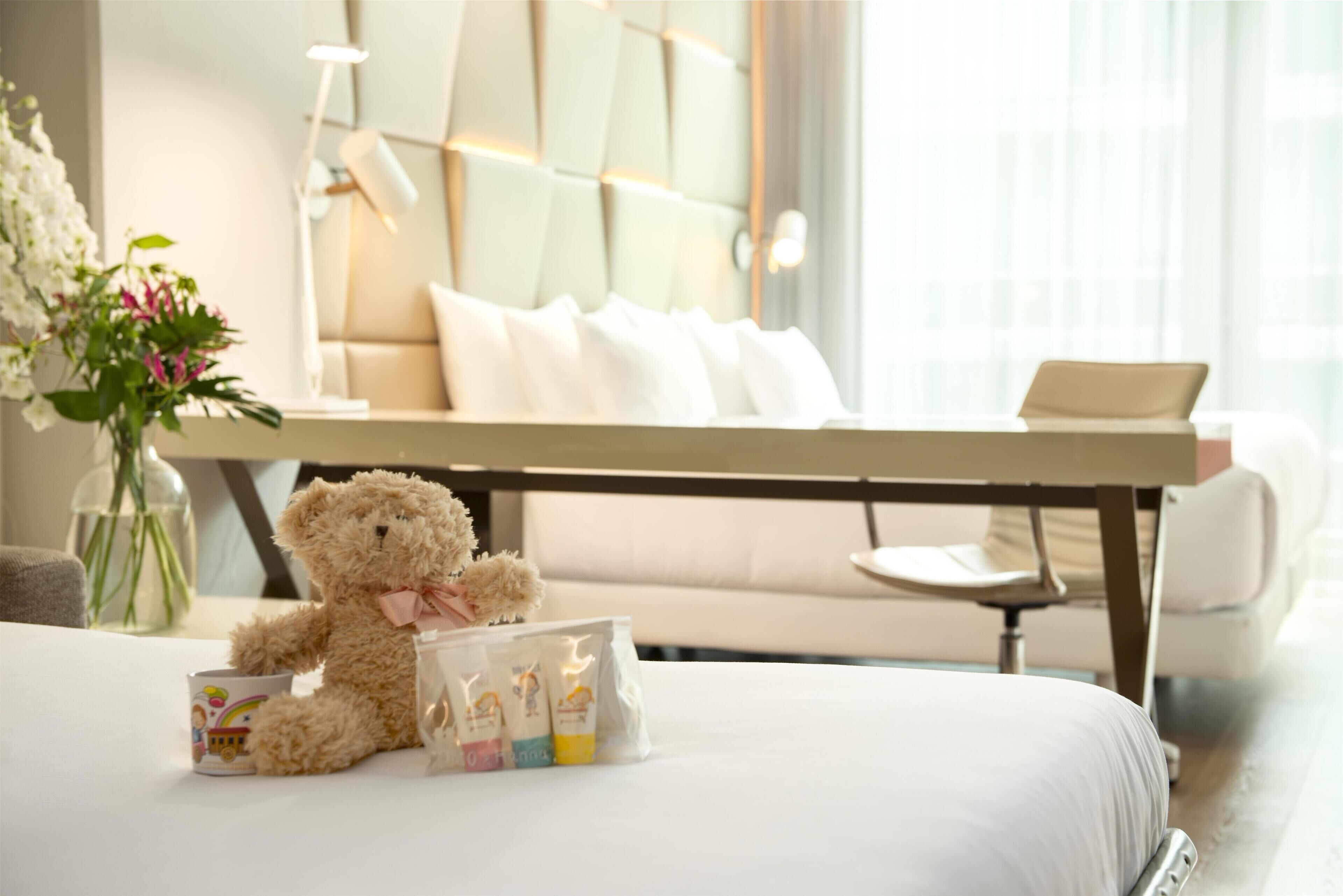 Ein ordentlich bezogenes Bett mit Teddybär, einer Blumenvase und einem Tisch mit Toilettenartikeln.