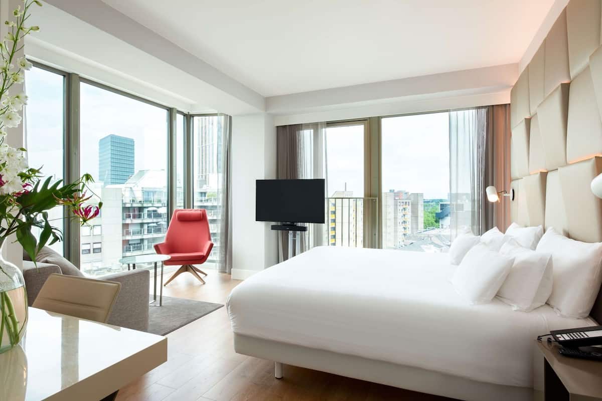 A hotel room with a large bed, a TV, a red chair, a desk, and a view of the city.