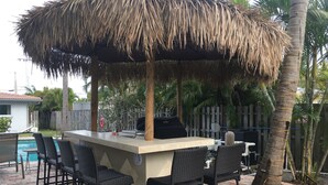 Un espace bar extérieur doté d’un toit de chaume, de tabourets de bar et d’une piscine en arrière-plan.