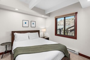 A hotel room with a large bed, a small table, a lamp, and a window with a view of a building.