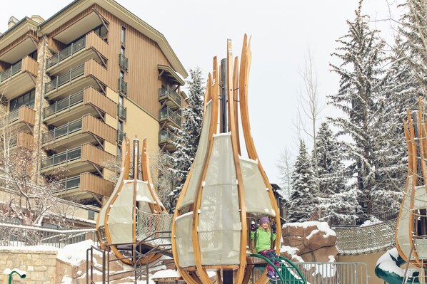 A ski lift with a person seated, surrounded by snow-covered trees and a multi-story building with balconies.