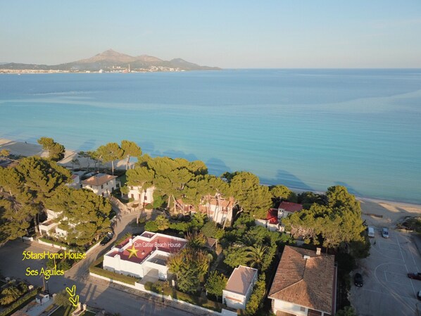 An aerial view of Can Cattar Beach House in Ses Aguiles, featuring a prominent star on the roof, surrounded by lush greenery and a clear view of the sea.