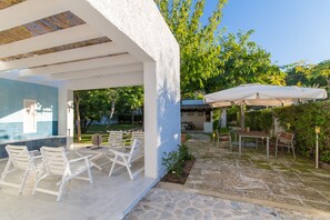 A covered outdoor seating area with white chairs and tables, stone flooring, and a view of the surrounding garden and trees.