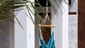 Un hamac est suspendu entre des piliers blancs dans un jardin, avec un palmier et des fleurs bleues au premier plan.