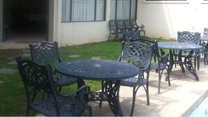 An outdoor patio area with a pool, featuring wrought iron tables and chairs, a covered seating area, and a building with large arched windows. - El Prado Hotel (Cochabamba)