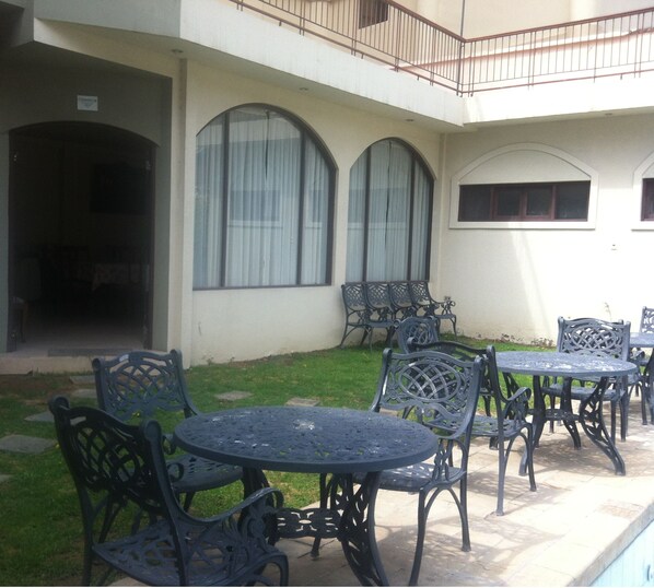 An outdoor patio area with a pool, featuring wrought iron tables and chairs, a covered seating area, and a building with large arched windows.