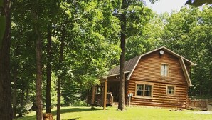 A rustic log cabin with a porch, surrounded by a grassy yard and trees.