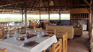 Un espacio de comedor al aire libre con mesas y sillas de madera, techo de paja y vistas al paisaje.