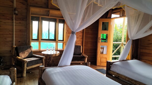 Habitación de cabaña de madera con dos camas, una silla, una mesa con botellas de agua y un ventanal grande.
