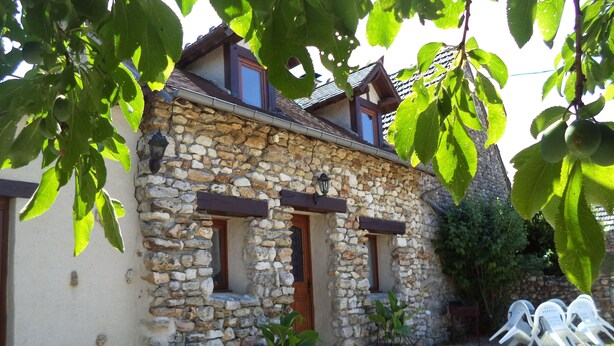 A stone building with a sloped roof, wooden shutters, and a small porch area with a chair and potted plants.