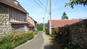 A narrow rural road flanked by stone buildings with red-tiled roofs and flowering plants.