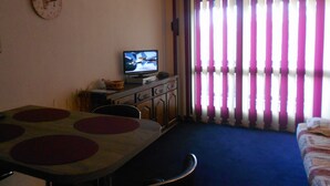 Une pièce comprenant un téléviseur posé sur un meuble, une table à manger avec des chaises, un lit et une fenêtre avec des rideaux à rayures.
