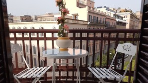 Een balkon met een ronde tafel en twee witte stoelen, een potplant en uitzicht op gebouwen.