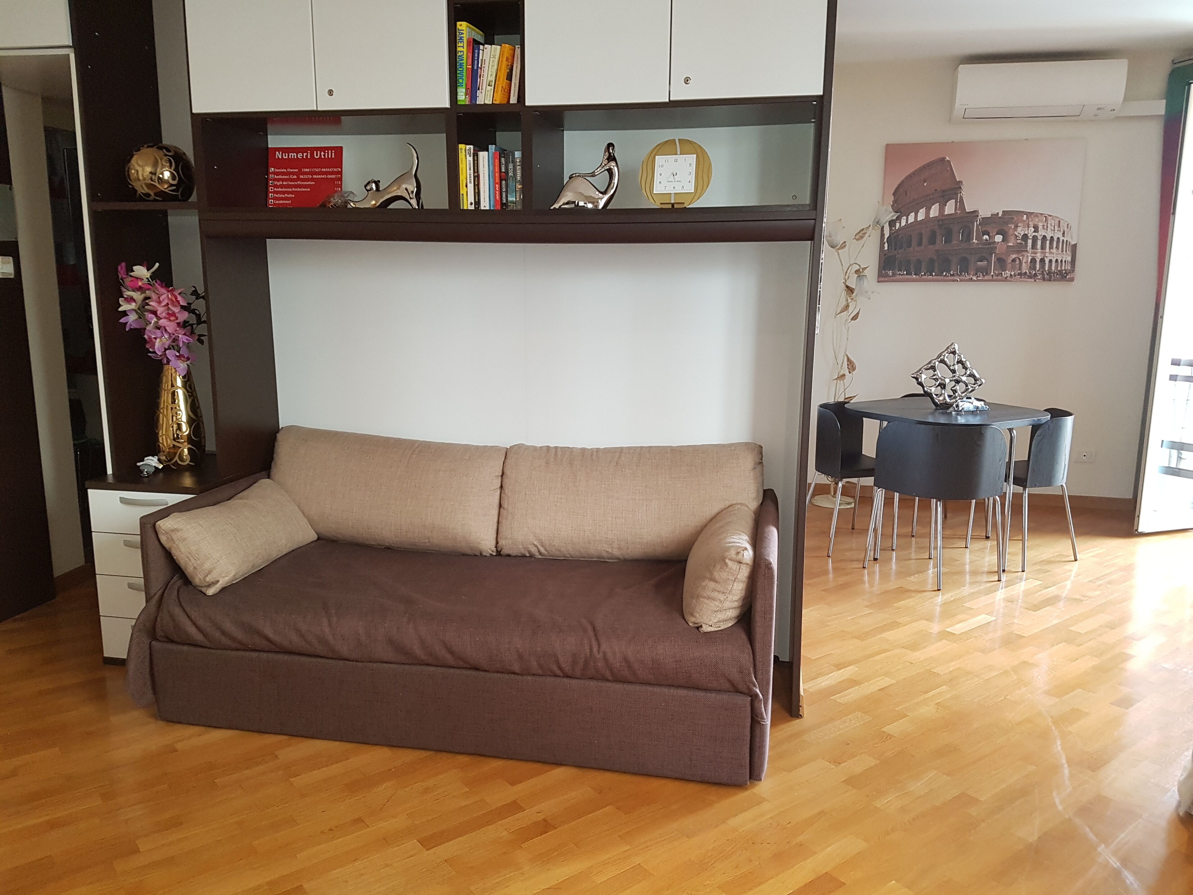 A living room with a brown sofa, a bookshelf with books and decorative items, a small table with chairs, and a wall-mounted air conditioner.