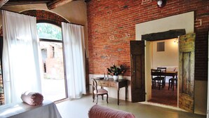 A room with a brick wall, a desk with a chair, a bed with a pink pillow, and a window with white curtains.