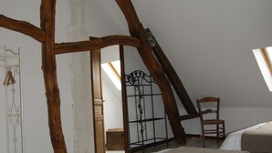 Een slaapkamer met een schuine balkenkap, zichtbare houten balken, een bed met een beige deken, een stoel en een metalen hek.