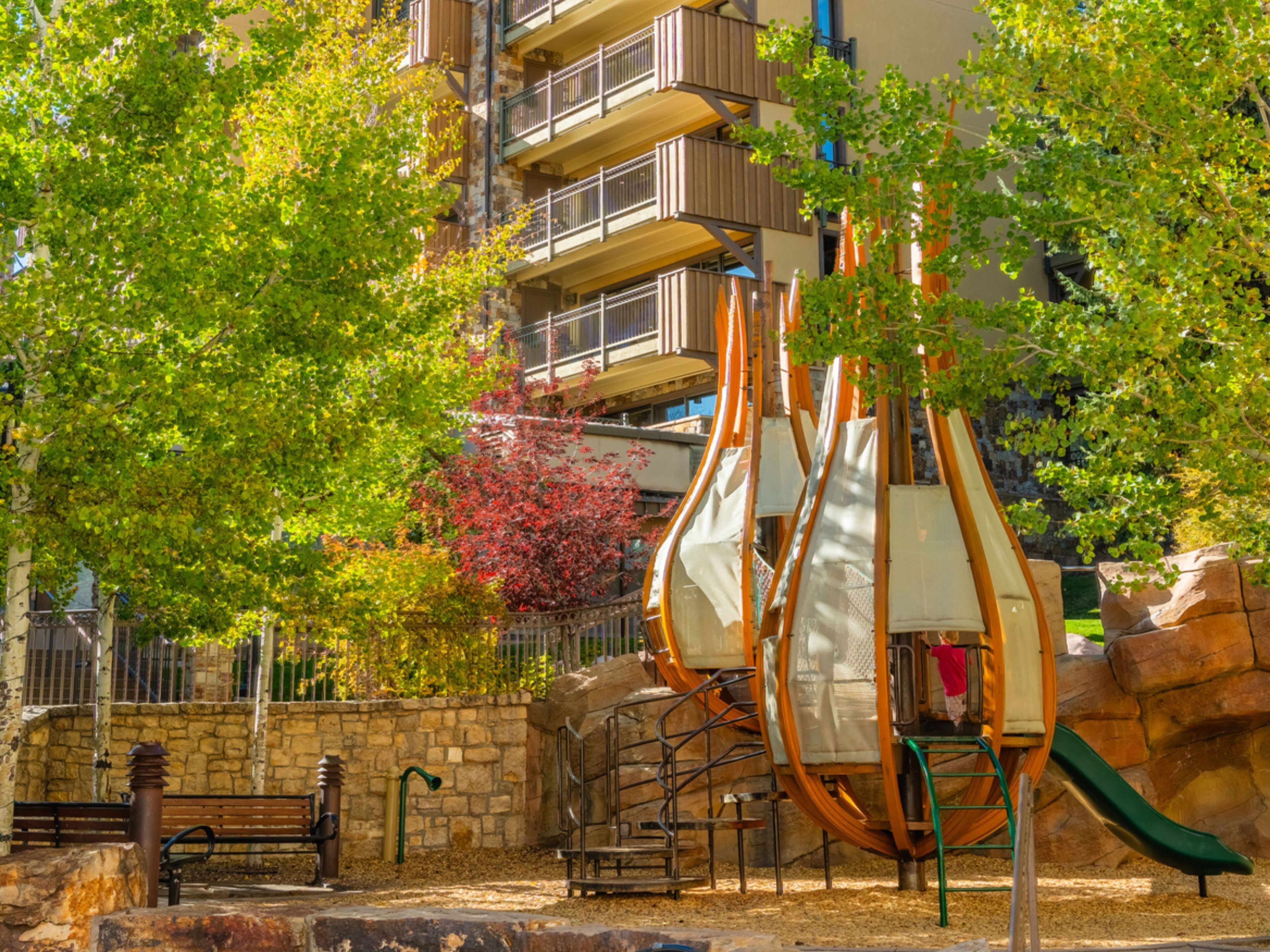 Un parc infantil amb tobogans i una banca, envoltat d'arbres i una paret de pedra.