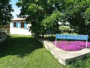 A sign reading "LES GENETS" in front of a building with blue shutters and a garden with purple flowers.