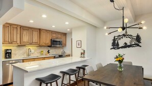 A modern kitchen with wooden cabinets, a central island with stools, a dining table, and a decorative wall piece. - Landmark 402 - 2 Bedroom Diamond (Vail)