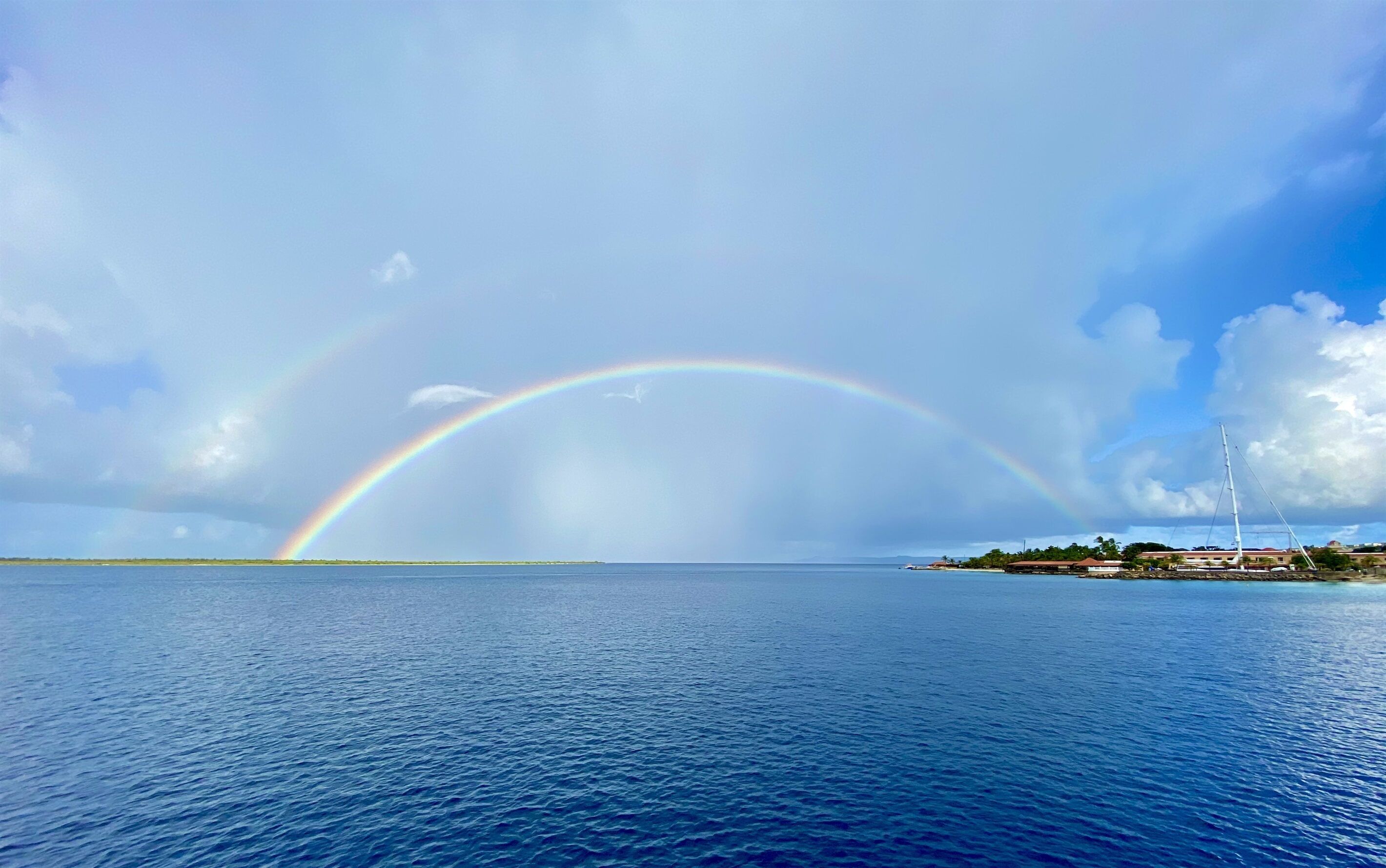 A coastal scene with a rainbow over the sea, a clear sky, and a distant shoreline with buildings.