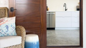A room with a wooden door, a woven sofa, a blue ottoman, a kitchen with white cabinets, and a hanging pendant light.