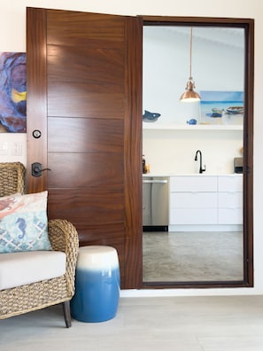 A room with a wooden door, a wicker sofa, a blue ottoman, a kitchen with white cabinets, and a hanging pendant light. - Kas di Alegria: Modern Studio with Tropical Charm, By the Sea, Walk to Downtown (Kralendijk)