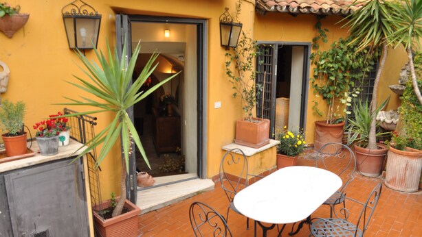 A courtyard with a table and chairs, potted plants, and a door leading to an interior space. - A Gorgeous Large Flat in Trastevere with a Beautiful Terrace (Roma)