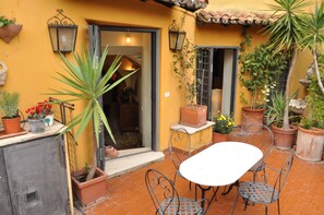 Um pátio com mesa e cadeiras, plantas em vasos e uma porta que dá para o interior.
