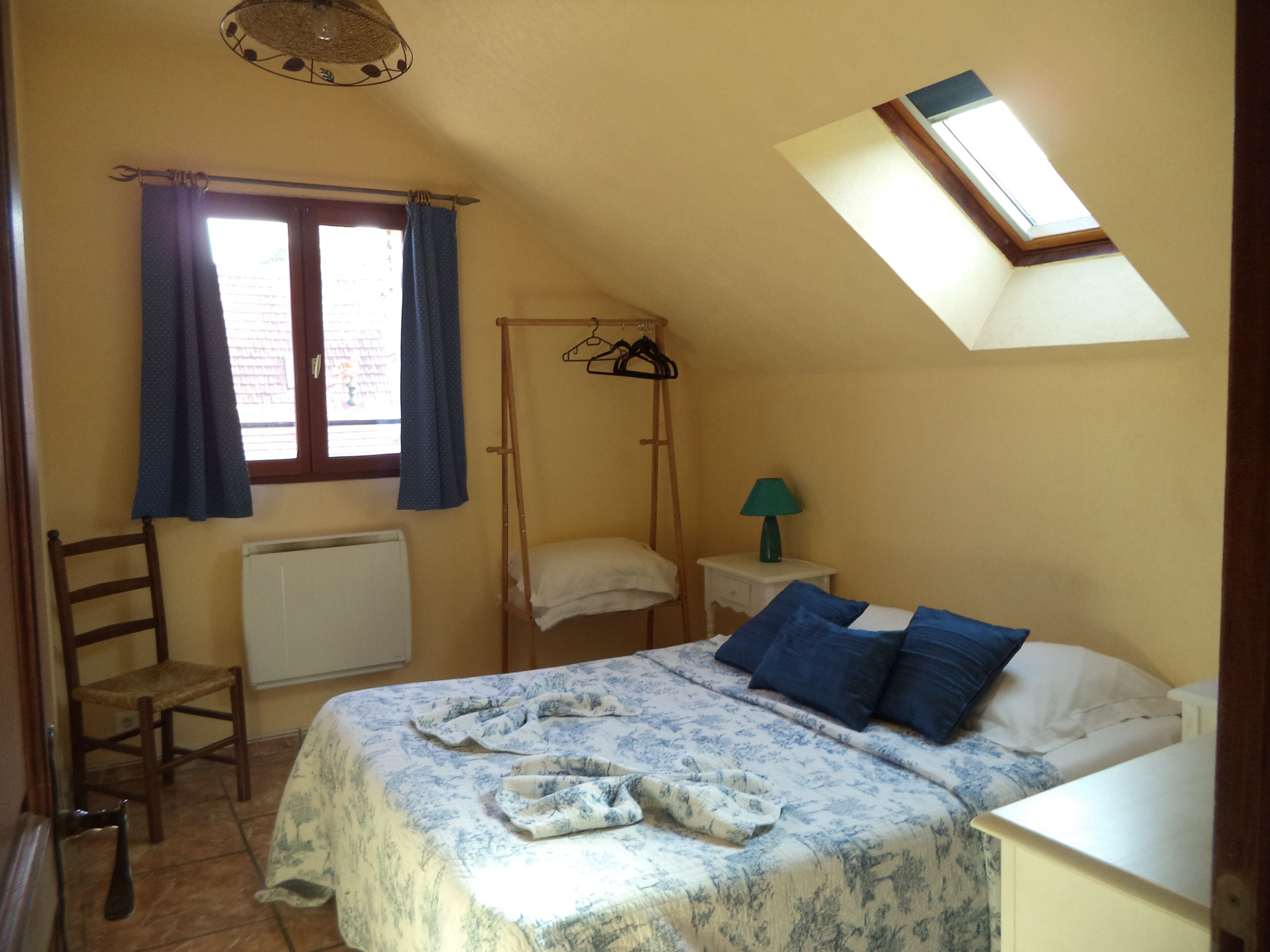 A bedroom with a sloped ceiling, a bed with a floral bedspread, a wooden chair, a radiator, a window with curtains, and a skylight.
