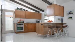 A modern kitchen with wooden cabinets, a built-in oven, a large island with bar stools, and a window overlooking a white fence. - LOVE ... and MISCHIEF in Historic Twin Palms (Palm Springs)