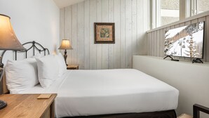 Un lit avec une literie blanche, une table de chevet en bois avec une lampe, un tableau encadré au mur et une fenêtre donnant sur un paysage enneigé.