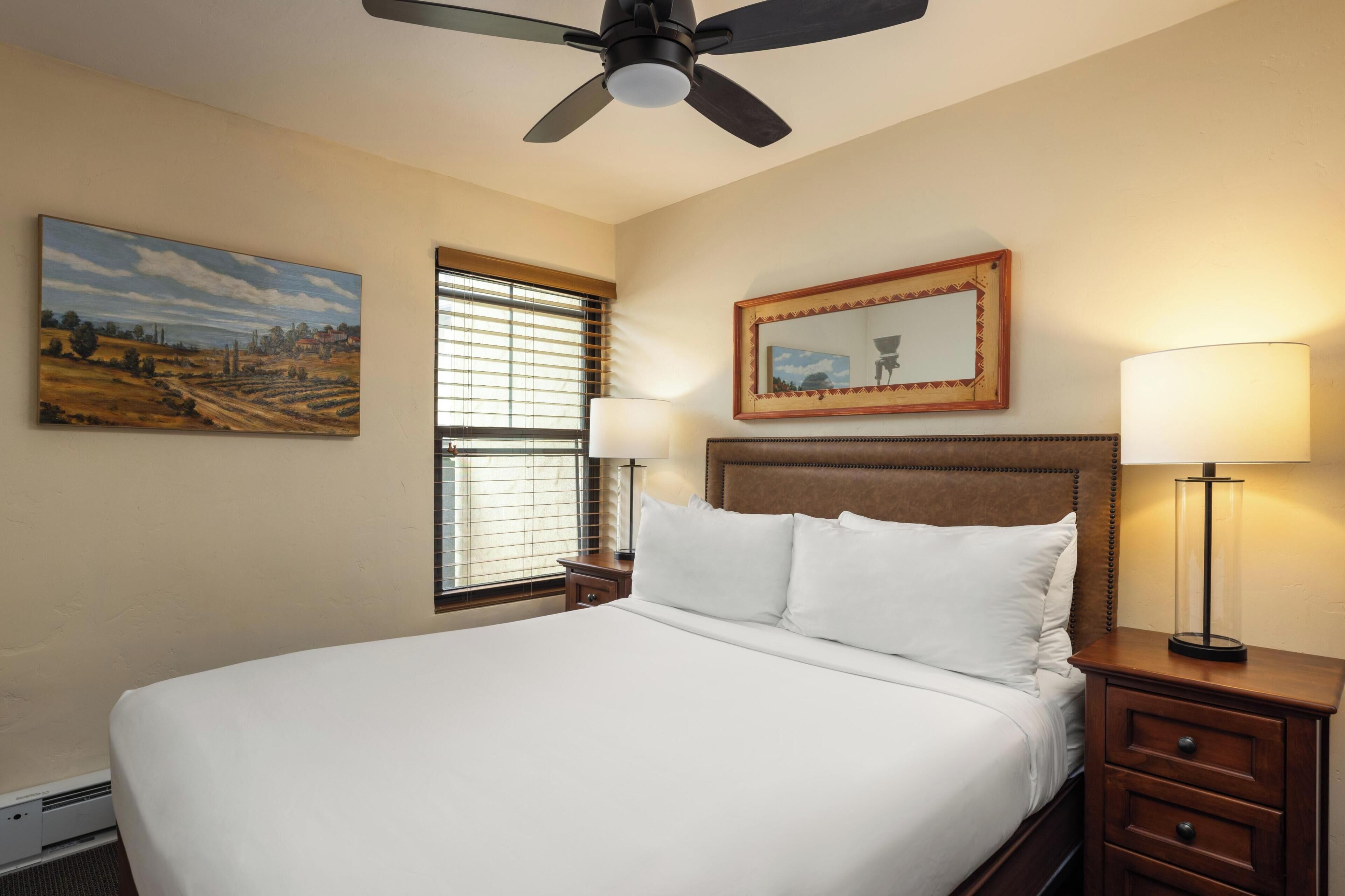 Habitación de hotel con una cama grande, dos mesitas de noche con lámparas, un ventilador de techo, un cuadro en la pared y una ventana con persianas.