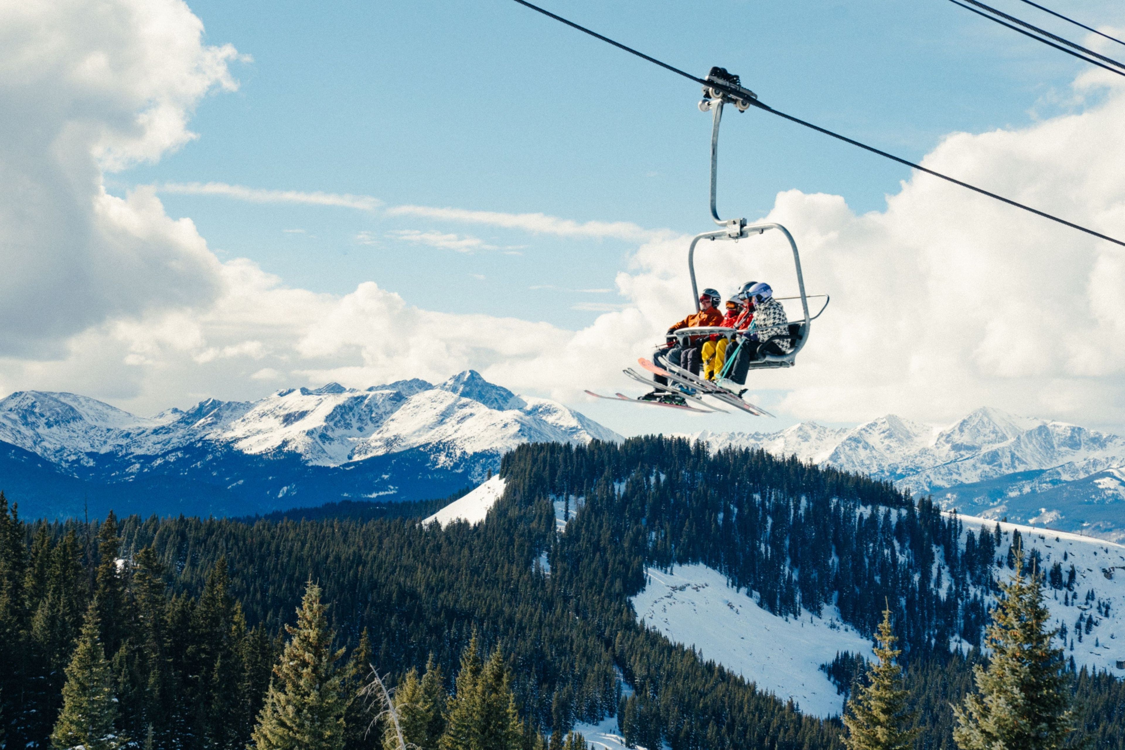 Een stoeltjeslift met skiërs die een besneeuwde berghelling beklimmen, omringd door dicht bos en besneeuwde bergtoppen.