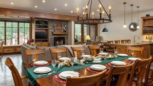 A dining area with a wooden table set for six, surrounded by wooden chairs. There is a fireplace with a stone surround, a television mounted on the wall, and a chandelier above the table.