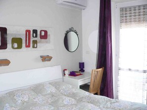 Ein Schlafzimmer mit Bett, Schreibtisch, Stuhl, Spiegel und einem Fenster mit Vorhängen.